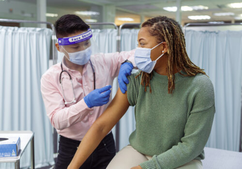 A young black woman wearing a protective face mask sits in a medical clinic as her male doctor of Asian descent prepares to give her a COVID-19 vaccination injection in the upper arm.