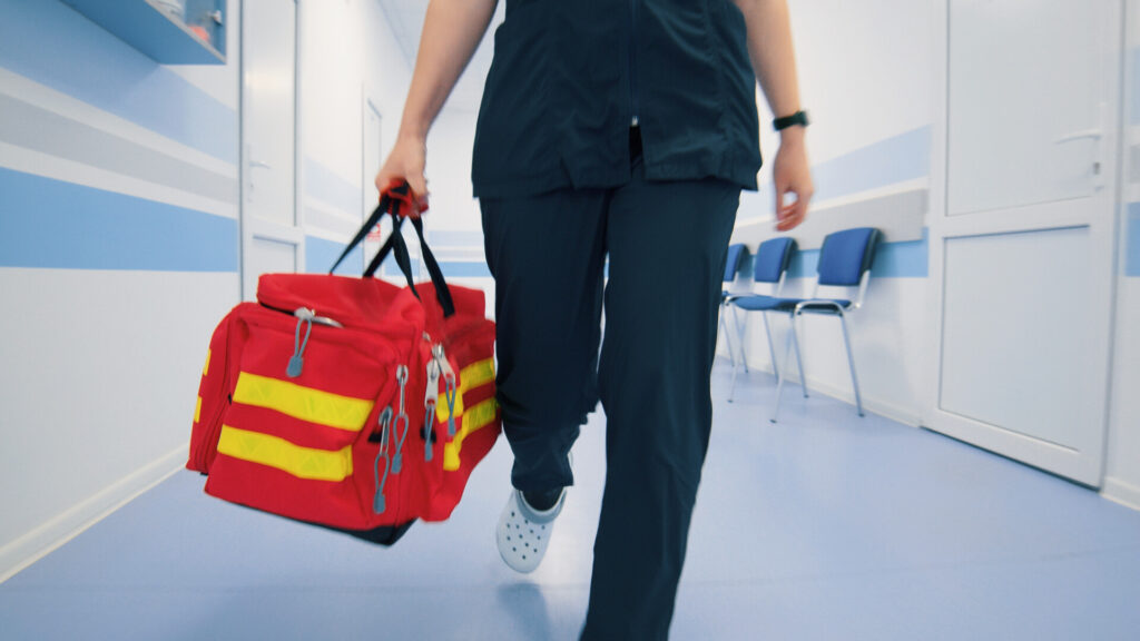 Paramedic walking through a clinic hallway, carrying a red emergency bag with yellow stripes, prepared for urgent care.