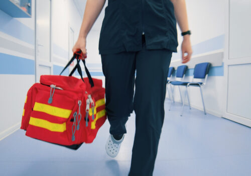 Paramedic walking through a clinic hallway, carrying a red emergency bag with yellow stripes, prepared for urgent care.