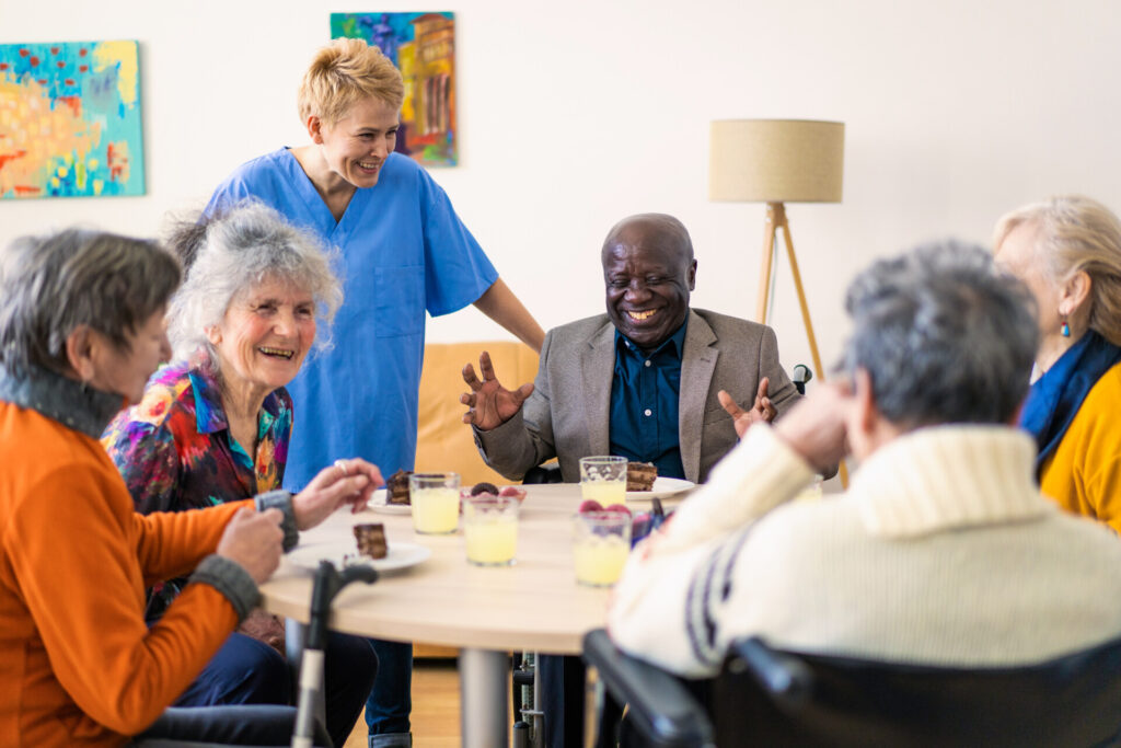 A cheerful nursing orderly is joining in the conversation with a group of pensioners sitting around a table having refreshments in a bright modern care home