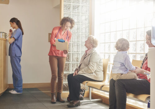 A female doctor stands next to a senior patient in her waiting room and casually chats through her factsheet . In the background a nurse stands at the reception desk whilst a mother and child play in the foreground waiting for their turn.
