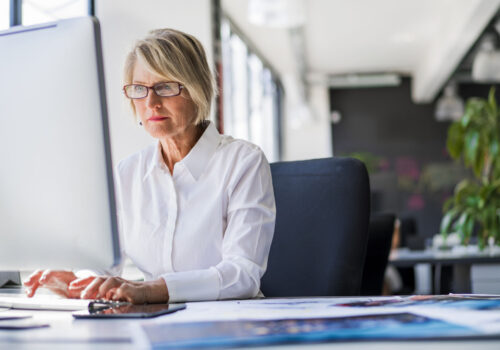 A photo of mature businesswoman using computer at desk. Concentrated female executive is working on desktop PC. Confident female professional is in formals. She is in brightly lit office.
