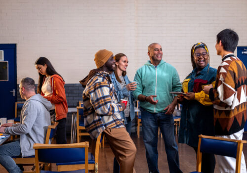 Three-quarter-length shot of a group of adults standing in a circle and talking to each other about their mental well-being. There is a staff member sitting to the left of them in the frame sitting next to a table. The community centre is located in Seaton Deleval in the North East of England.